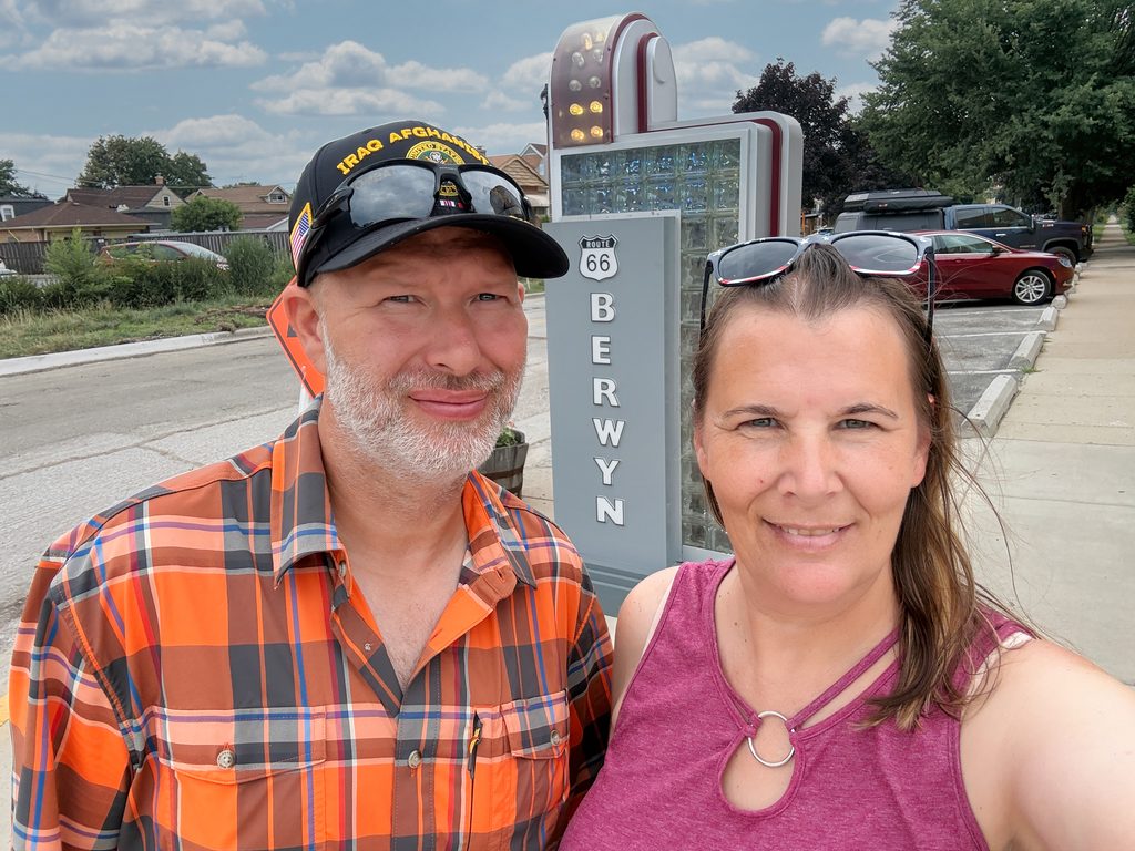 man and woman standing in front of an old fashioned sign with a route 66 shield and the word Berwyn.