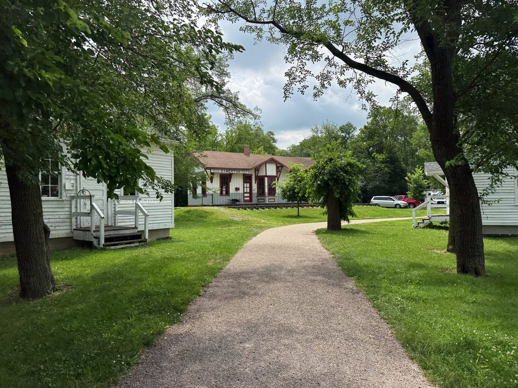 Walking path leading through a grassy area lined with trees and white historic buildings, ending at the Symerton railroad depot in the background under a partly cloudy sky.