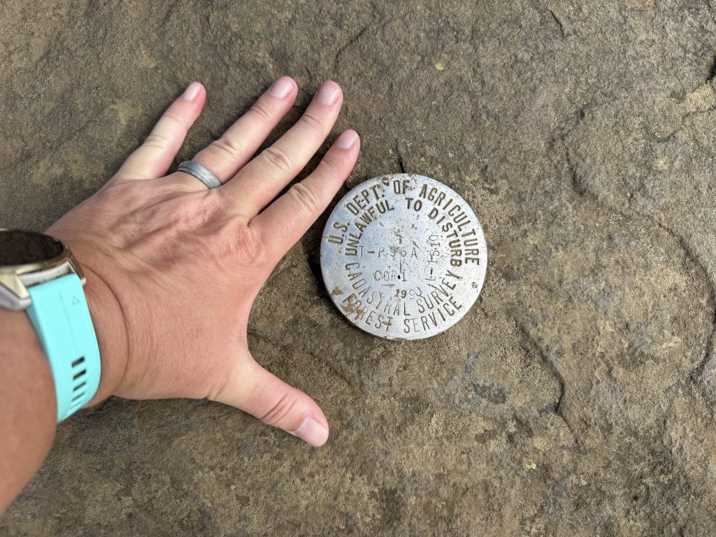 survey marker with my hand next to it at the top of Grassy Ridge
