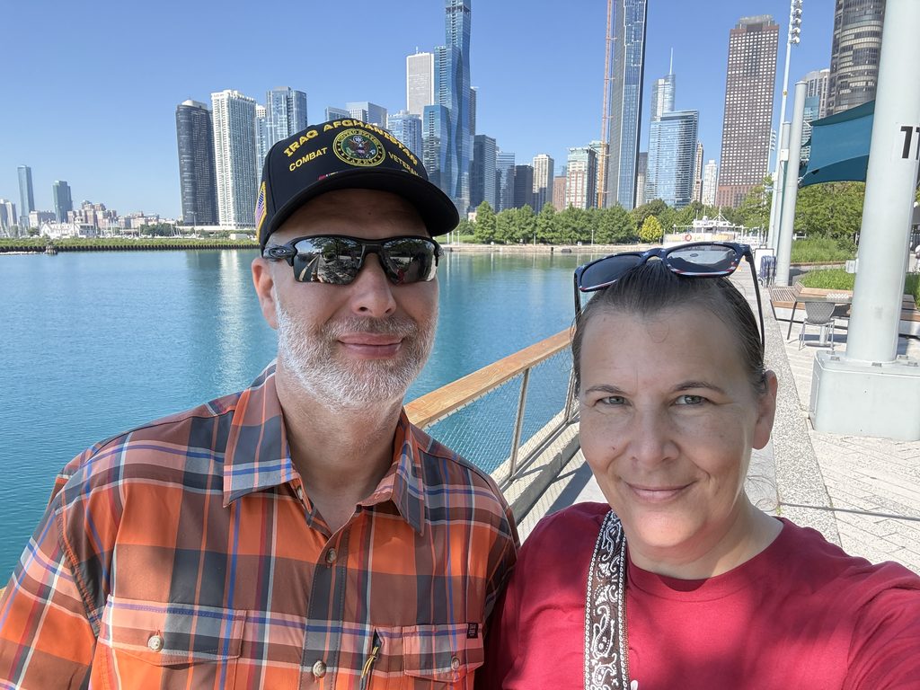 A couple in a plaid shirt and red Route 66 shirt poses in front of Chicago’s skyline on Navy Pier with trees, the Chicago skyscrapers, and bay visible behind them.