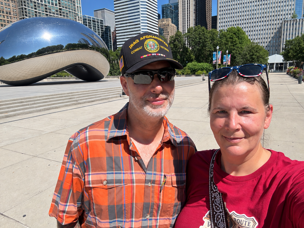 A couple in a plaid shirt and red Route 66 shirt poses in front of Chicago’s Cloud Gate in Millennium Park, with the empty plaza, trees, and Chicago skyscrapers visible behind them.