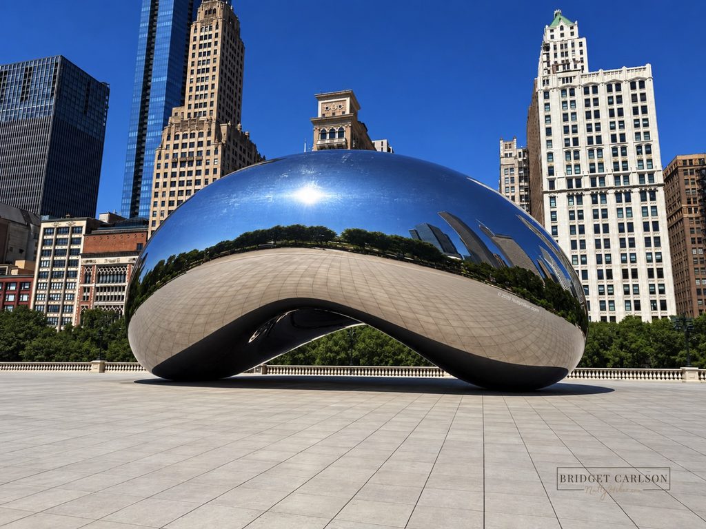 Cloud Gate, the reflective Bean sculpture in Chicago’s Millennium Park, surrounded by skyscrapers and trees under a clear blue sky, with the sculpture reflecting the park and skyline.