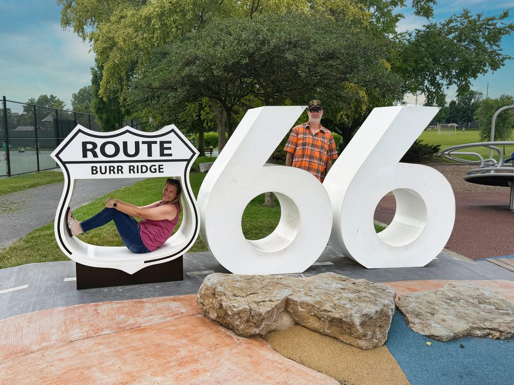 A woman sits playfully inside a large decorative Route Burr Ridge sign shaped like a highway shield, next to oversized white “66” numerals forming a “Route 66” display. A man in an orange plaid shirt and cap stands behind the numerals. The scene is set in a park with green grass, trees, rocks in the foreground, a playground area, and tennis courts visible in the background.