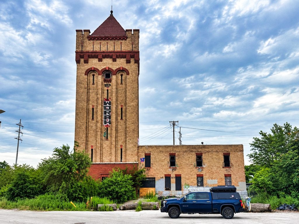 Straight-on view of the abandoned Hawthorne Works tower in Cicero, Illinois, featuring a tall tan-brick tower with red architectural details and a vertical sign, attached to a graffiti-covered brick building. Overgrown vegetation, utility poles, and a dark blue pickup truck with a camper shell sit in the foreground beneath an overcast sky.