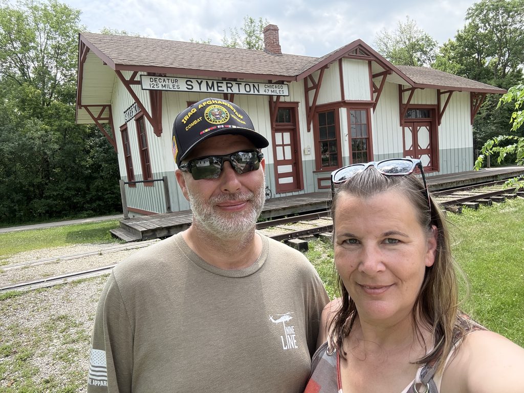 Two people posing for a selfie in front of the historic Symerton railroad depot, with its white and red wooden exterior and railway tracks visible beside them.