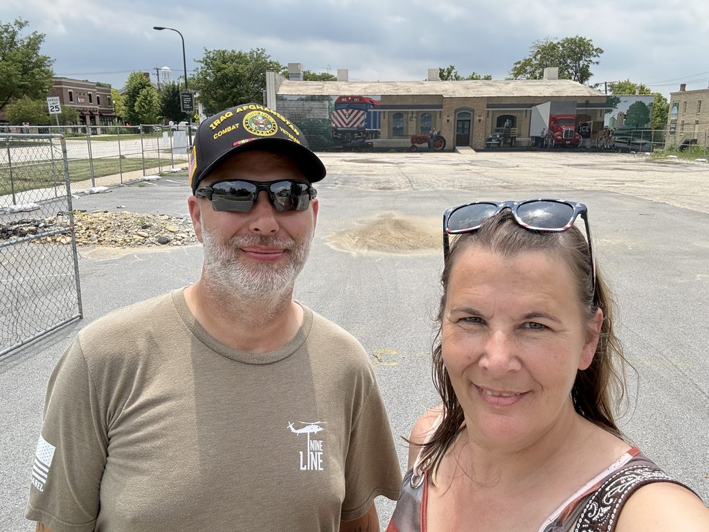 Two people standing in a paved lot with construction fencing, posing for a selfie. Behind them is a building with a colorful mural depicting vintage vehicles and local scenes, under a partly cloudy sky.