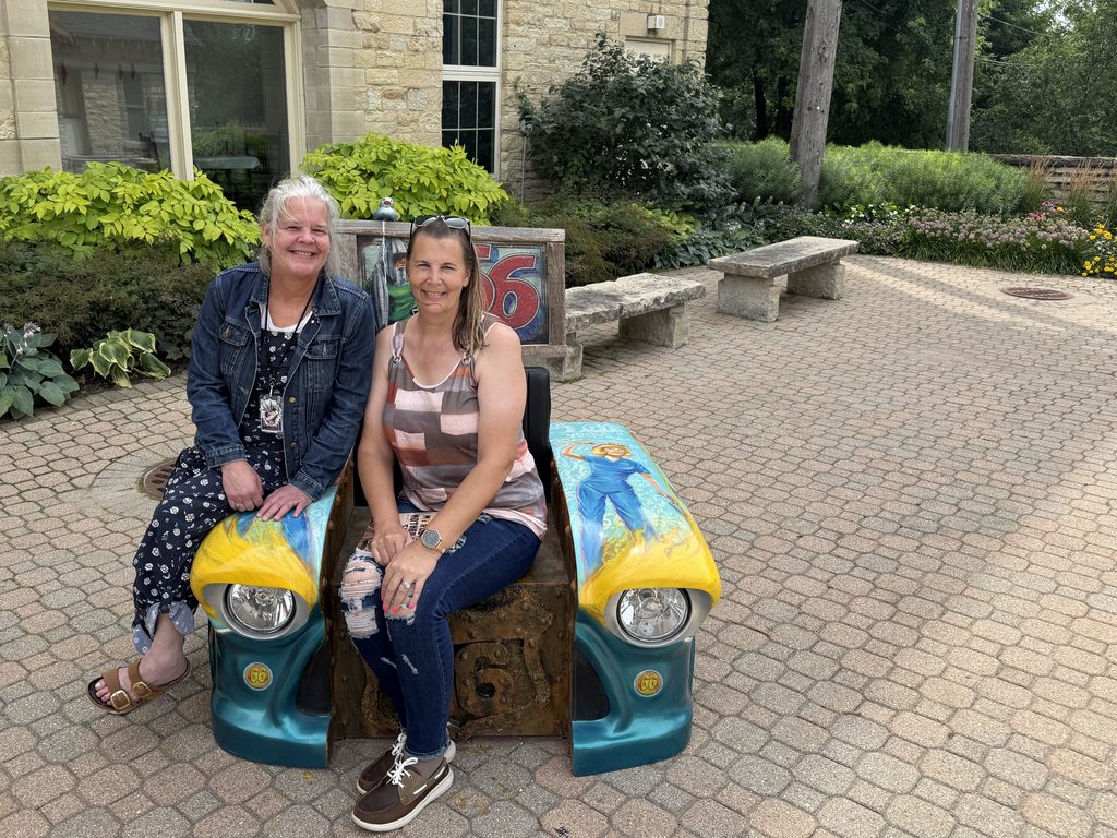 Two women sitting and smiling on a colorful bench made from vintage car parts, with painted headlights and Route 66 artwork, outside a stone building surrounded by landscaped greenery and brick pavers.