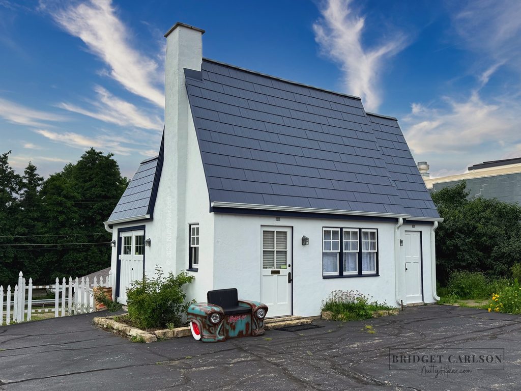 A small, white historic building with a steep, dark slate roof and a central chimney, surrounded by greenery and a white picket fence. In front of the building is a bench made from an old car front end, all set against a bright blue sky with wispy clouds.