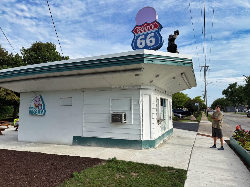 Small white ice cream stand with a teal roof and a large Route 66 sign on top, along with a black statue of a man sitting beside the sign. A man stands outside the building, which has a "Rich & Creamy" ice cream sign, on a sunny day with a partly cloudy sky.
