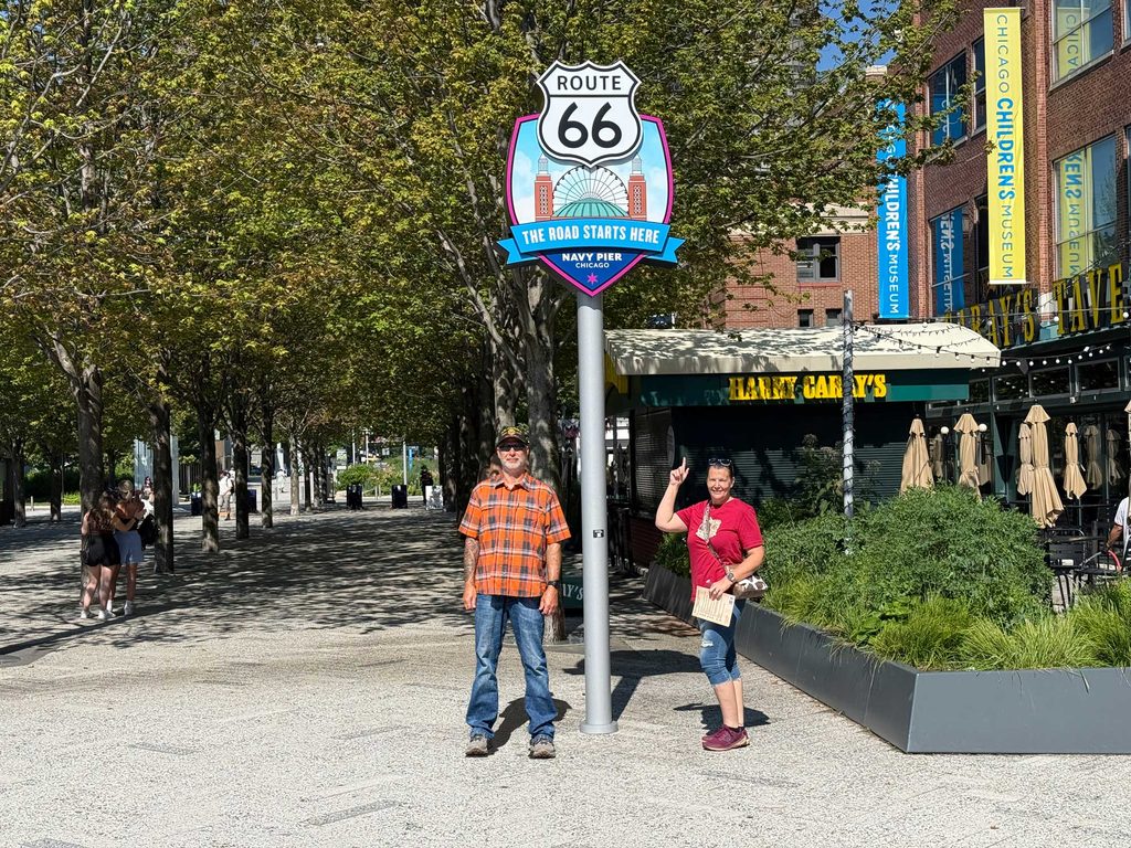 Route 66 sign at Navy Pier in Chicago with a man in an orange plaid shirt and a woman in a red shirt standing beside the sign on a tree-lined pedestrian walkway.