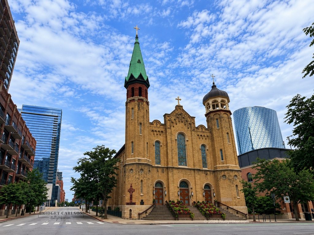 St. Patrick’s Catholic Church in Chicago, featuring a historic tan-brick façade, twin towers with green and gray spires, stained-glass windows, and flower-filled steps, framed by downtown buildings and trees beneath a blue, cloud-filled sky.