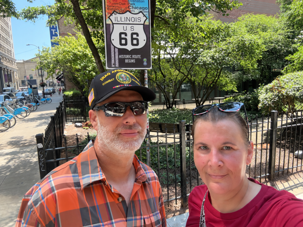 Couple posing in front of the Historic Route 66 Begins sign in Chicago, Illinois, with the Route 66 sign visible above them on a sunny tree-lined city street.