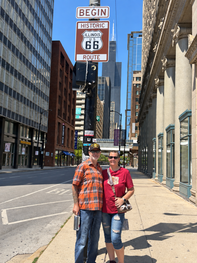 A couple stands on a Chicago sidewalk beneath a “BEGIN HISTORIC ILLINOIS US 66 ROUTE” sign, with tall downtown buildings, a quiet street, and the Willis Tower visible in the background on a sunny day.