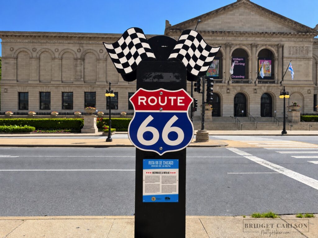 A clean Route 66 sign with checkered flags and a Spanish informational plaque stands in front of the Art Institute of Chicago, with the museum’s ornate stone façade, landscaped entrance, crosswalk, and city street visible on a sunny day.