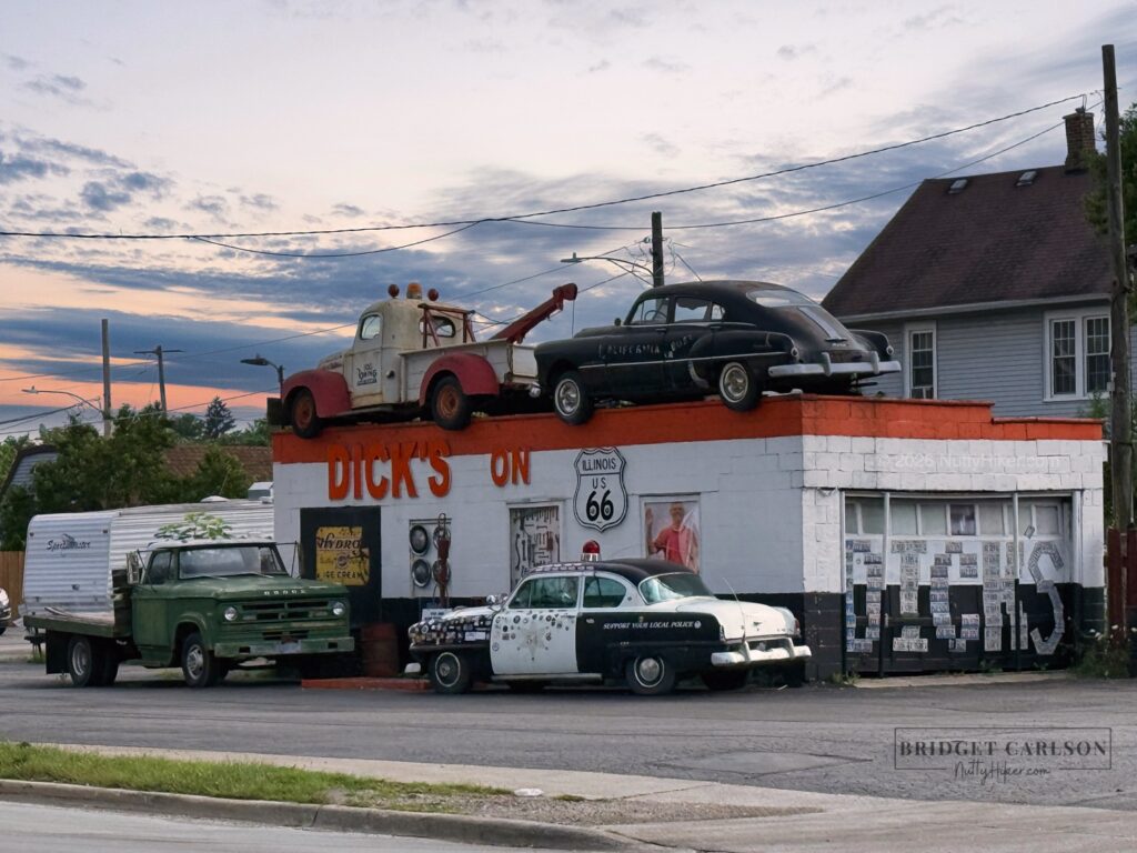 Vintage roadside building labeled "Dick's on Route 66," with an old tow truck and black car displayed on the roof, and classic vehicles, including a green truck and black-and-white police car, parked in front. The building has an Illinois Route 66 sign and retro signage, set against a neighborhood backdrop at sunset.