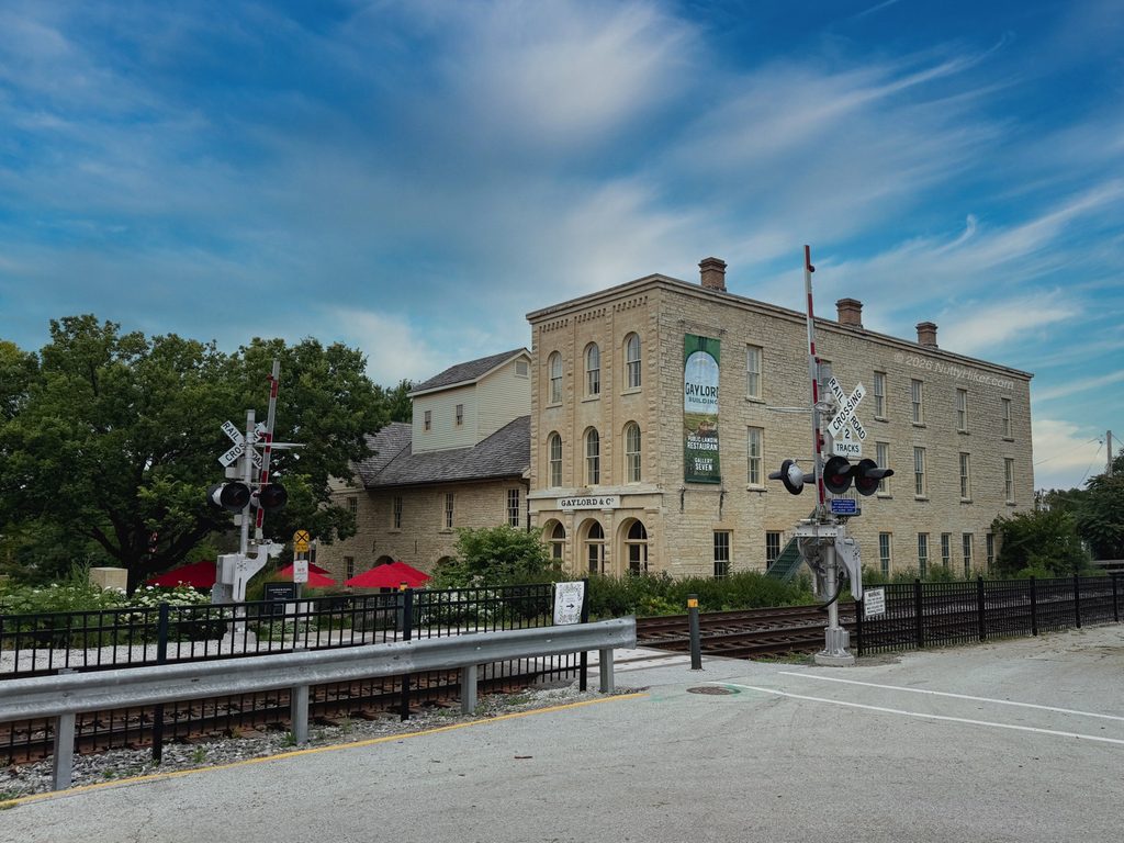 Exterior view of the historic Gaylord Building in Lockport, Illinois, with its light limestone facade, visible railway crossing, and lush greenery under a bright blue sky.