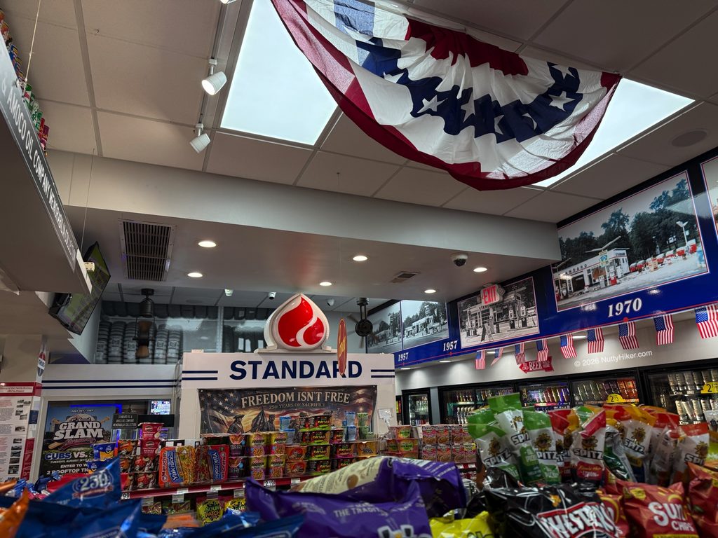Interior of a convenience store decorated with patriotic bunting, a large skylight, and a red Standard Oil logo sign. The store displays a variety of snacks in the foreground, with framed historical photos and small American flags lining the top of the refrigerated section. A banner reads "FREEDOM ISN'T FREE."