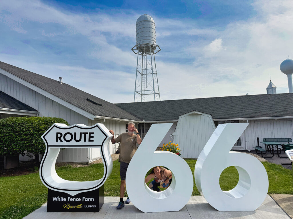 Two people posing with a large white Route 66 sign at White Fence Farm in Romeoville, Illinois, with a water tower and farm buildings in the background. One person stands behind the sign and the other crouches inside the hollow “6.”