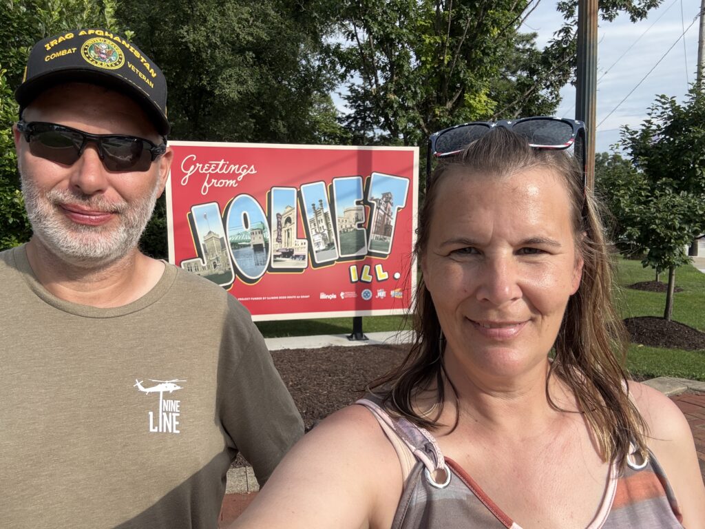 Two people smiling and taking a selfie in front of a large red "Greetings from Joliet, ILL." sign, with trees and landscaping in the background.