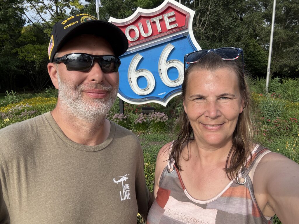 Two people smiling for a selfie in front of a large vintage-style Route 66 sign, surrounded by colorful flowers and greenery.