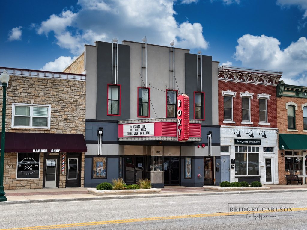 The historic Roxy Theater in a small-town downtown, featuring its vertical red sign and marquee advertising a free live bluegrass jam. The theater is flanked by a barber shop, Edward Jones office, and neighboring storefronts along an empty street with double yellow lines.