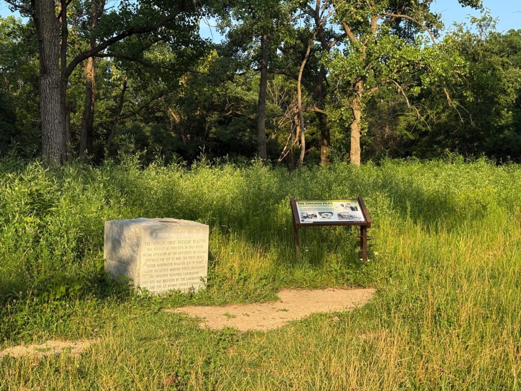 Stone marker and informational sign standing side-by-side in a small clearing surrounded by tall grass and trees at Red Gate Woods.