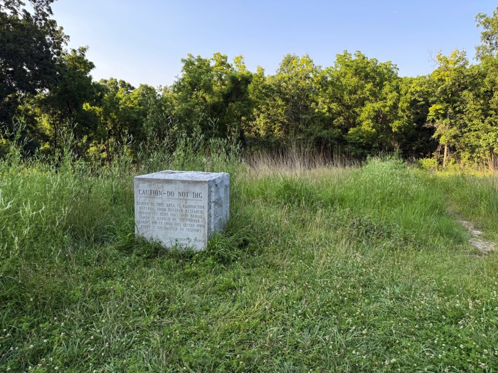 Concrete marker in a grassy field that reads "Caution—Do Not Dig," warning of buried radioactive material, surrounded by green vegetation and trees in the background.