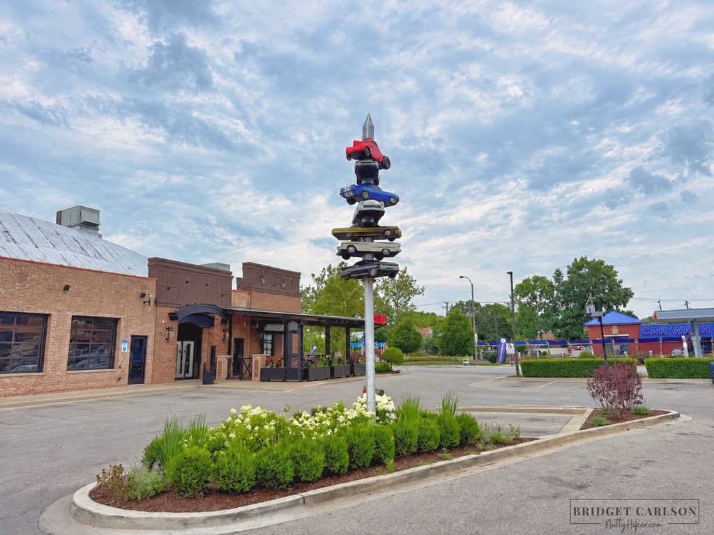 A parking lot with a unique sculpture made of stacked, colorful toy cars mounted on a tall pole as its centerpiece. The sculpture sits in a landscaped island with green shrubs and white flowers. In the background, there is a brick building with large windows and a metal roof, and to the right, a blue and red car wash building. The sky is partly cloudy, and the area appears quiet and well-maintained.