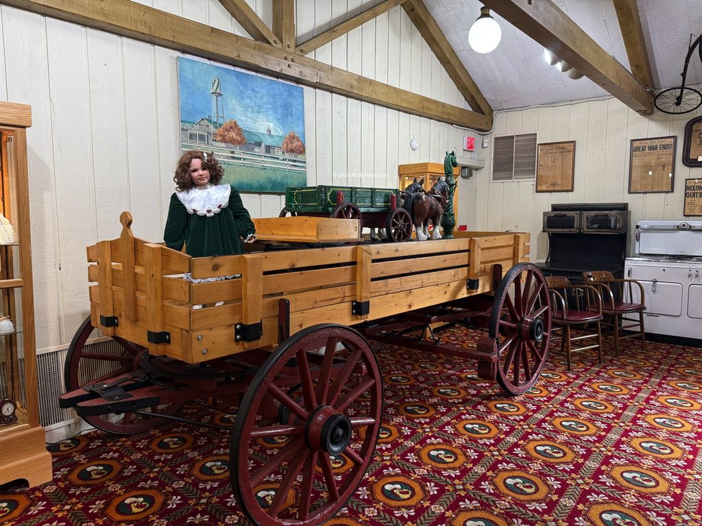 Indoor scene featuring a large wooden wagon with red wheels, displayed on a patterned carpet. A life-size doll in a green dress and white lace collar stands in the wagon, with miniature horse and wagon figurines beside her. The room has vintage decor, an old stove, framed pictures, and wooden beams on the ceiling.